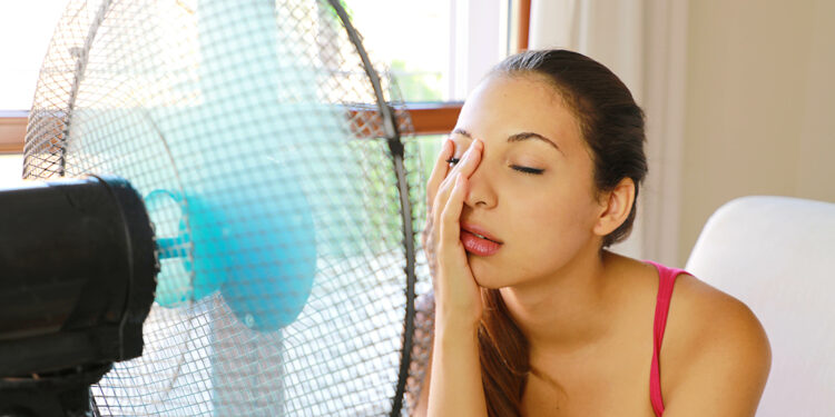 Young woman suffering a heat wave using a fan sitting on a couch in the living room at home.